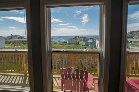 A balcony with a wooden railing, a red chair, and a view of a coastal area with houses and the sea.. Whistler's View