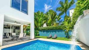 Una piscina con agua cristalina azul, rodeada por un edificio blanco, un patio con sillas y abundante vegetación.