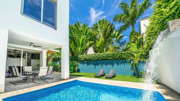 A swimming pool with a clear blue water, surrounded by a white building, a patio with chairs, and lush greenery. - The Castle II - Modern Villa with Private Pool at a Safe Gated Community (Cancún)