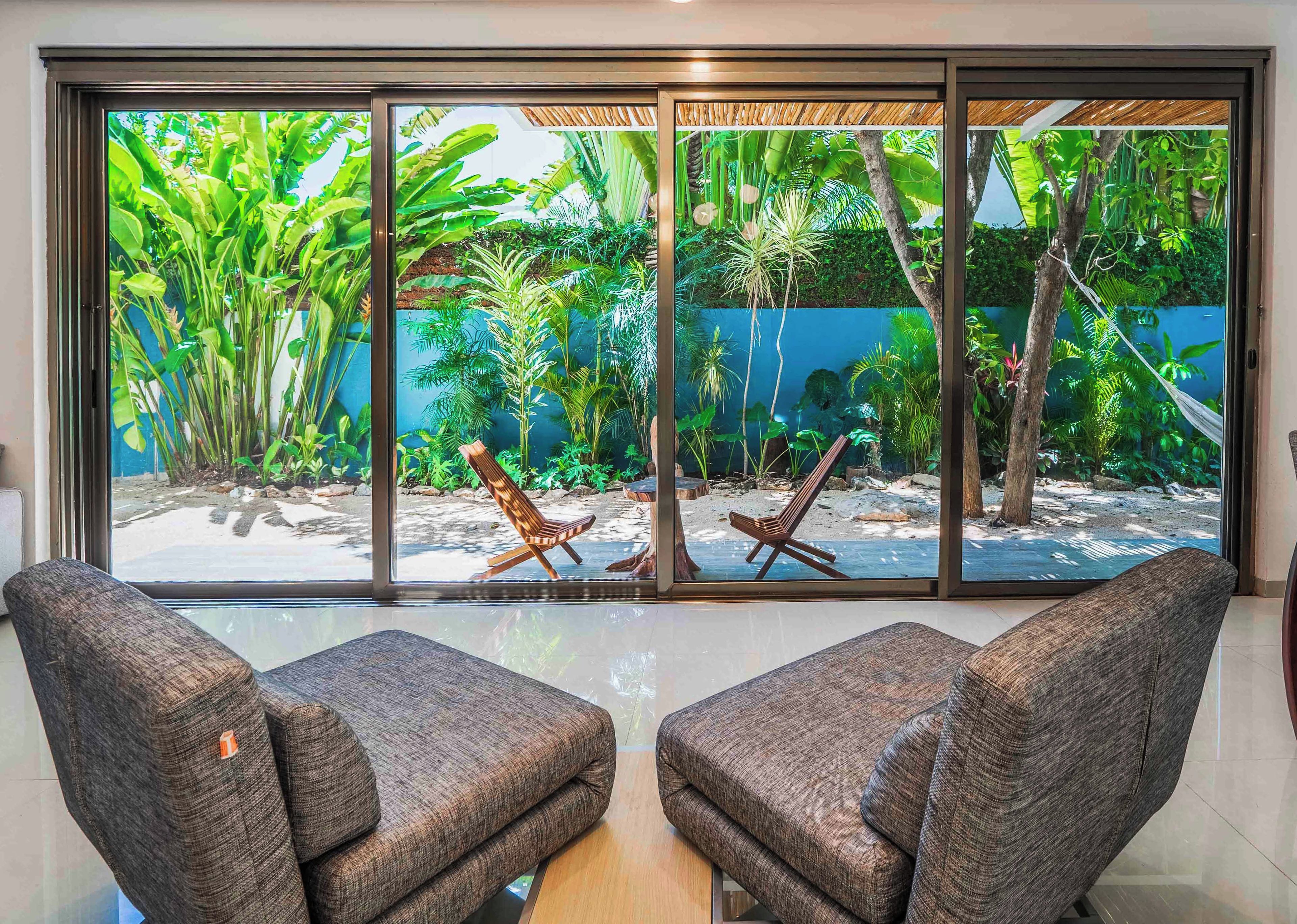 A room with a large window overlooking a tropical garden with chairs.
