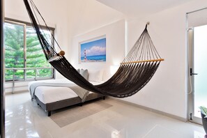 A modern bedroom with a hammock bed, a large window with a view of greenery, and a painting of a lighthouse on the wall.