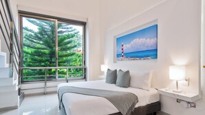 A modern bedroom with a large bed, a painting of a beach scene, a window with a view of trees, and a nightstand with a lamp. - The Castle II - Modern Villa with Private Pool at a Safe Gated Community (Cancún)