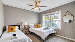 A hotel room with two beds, a ceiling fan, a window with a view, and a round mirror on the wall. - Oakley Square 2BR Condo-Walkable with Free Parking (Cincinnati)