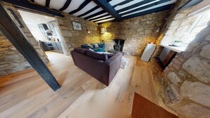 Un intérieur rustique avec des murs en pierre apparentes, un plancher en bois, un canapé, une cheminée et une cuisine en arrière-plan.