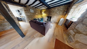 A rustic interior with exposed stone walls, a wooden floor, a sofa, a fireplace, and a kitchen in the background.