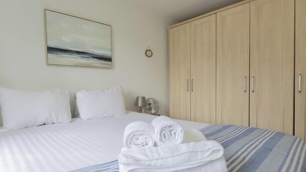 A neatly made bed with white linens and rolled towels, a bedside table with a lamp, and a large wardrobe. - Bridge View | Oxford Holiday Let | 1 Bed & Sleeps 2 (Oxford)