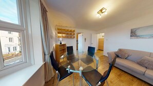A modern living room with a glass dining table, a sofa, and a view of the street through a window. - Bridge View | Oxford Holiday Let | 1 Bed & Sleeps 2 (Oxford)