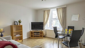 A living room with a sofa, TV, wooden bookshelf, and a dining area with chairs and a table. - Bridge View | Oxford Holiday Let | 1 Bed & Sleeps 2 (Oxford)