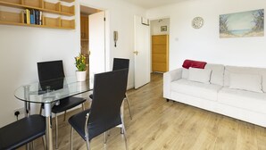 A modern living room with a glass dining table, black chairs, a white sofa, wooden shelves, and a painting on the wall. - Bridge View | Oxford Holiday Let | 1 Bed & Sleeps 2 (Oxford)