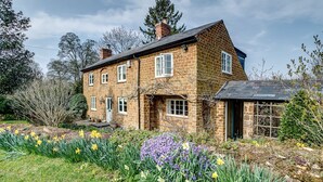 Een stenen huis met schuin dak, meerdere ramen en een tuin met narcissen en paarse bloemen.