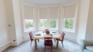 Een eetkamer met een ronde tafel, zes stoelen en een groot raam met jaloezieën.