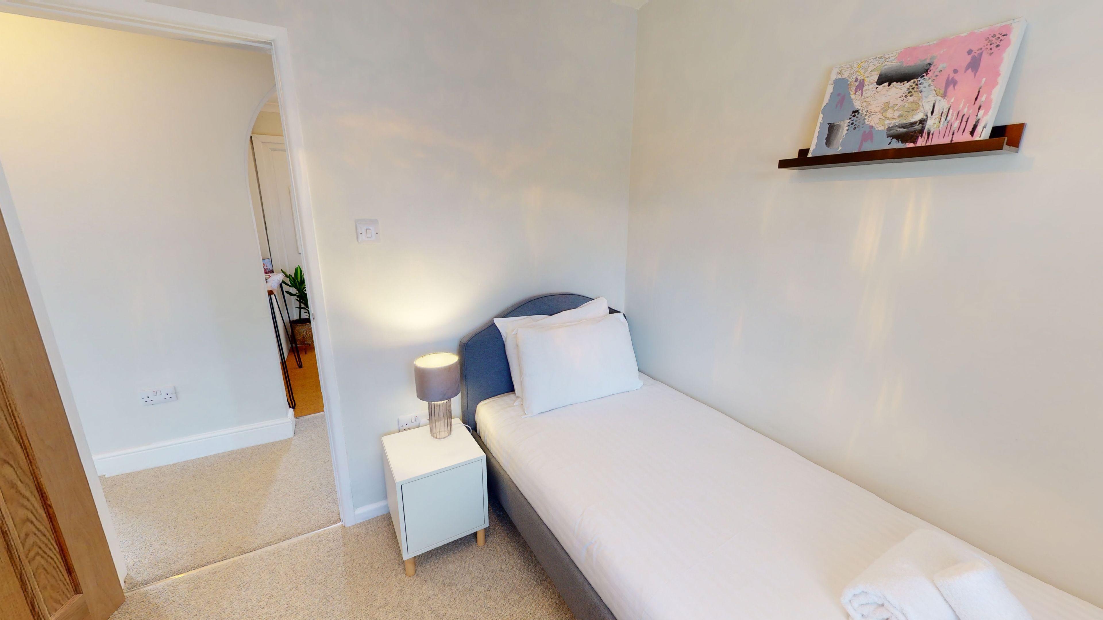 A small, neatly arranged bedroom with a single bed, a bedside table, a lamp, and a wall-mounted shelf with a painting.