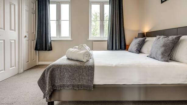 Un dormitorio con cama, dos ventanas con cortinas, una mesita de noche con lámpara y un cuadro enmarcado en la pared.