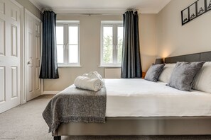 Ein Schlafzimmer mit einem Bett, zwei Fenstern mit Vorhängen, einem Nachttisch mit Lampe und einem gerahmten Bild an der Wand.