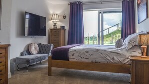 Une chambre à coucher avec un lit, une télévision, une commode, une chaise et une fenêtre avec des rideaux.
