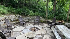 A stone patio with Adirondack chairs and a picnic table surrounded by a natural forest setting. - #4 Cabin, cottages, fishing, Hunting, Trails, nature, creek, ponds, lakes  (Oneida)