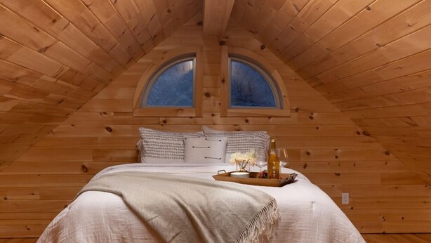 A cozy attic bedroom with a wooden ceiling, two skylights, a large bed with a beige blanket, pillows, and a tray with a bottle and flowers. - #4 Cabin, cottages, fishing, Hunting, Trails, nature, creek, ponds, lakes  (Oneida)