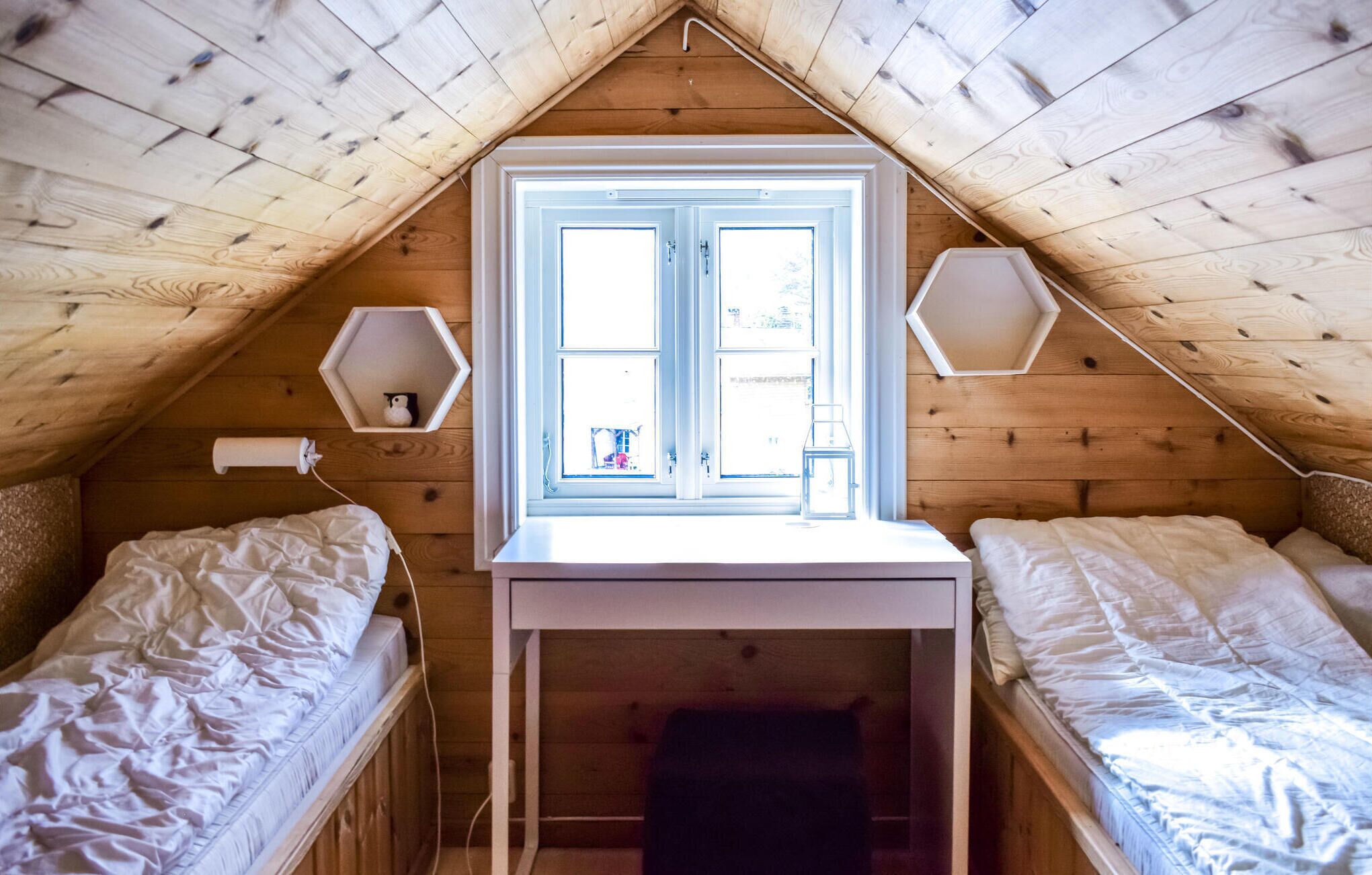 Une petite chambre mansardée avec deux lits simples, une fenêtre et deux appliques murales hexagonales.