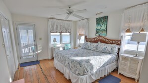 A bedroom with a bed, a ceiling fan, a window with curtains, a nightstand, and a painting on the wall. - High Tide - Beachfront Home in "Arrogantly Shabby" Pawleys Island, SC (Pawleys Island)