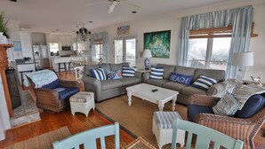 A spacious living room with a grey sofa, wicker chairs, and a coffee table. A kitchen is visible in the background. - High Tide - Beachfront Home in "Arrogantly Shabby" Pawleys Island, SC (Pawleys Island)