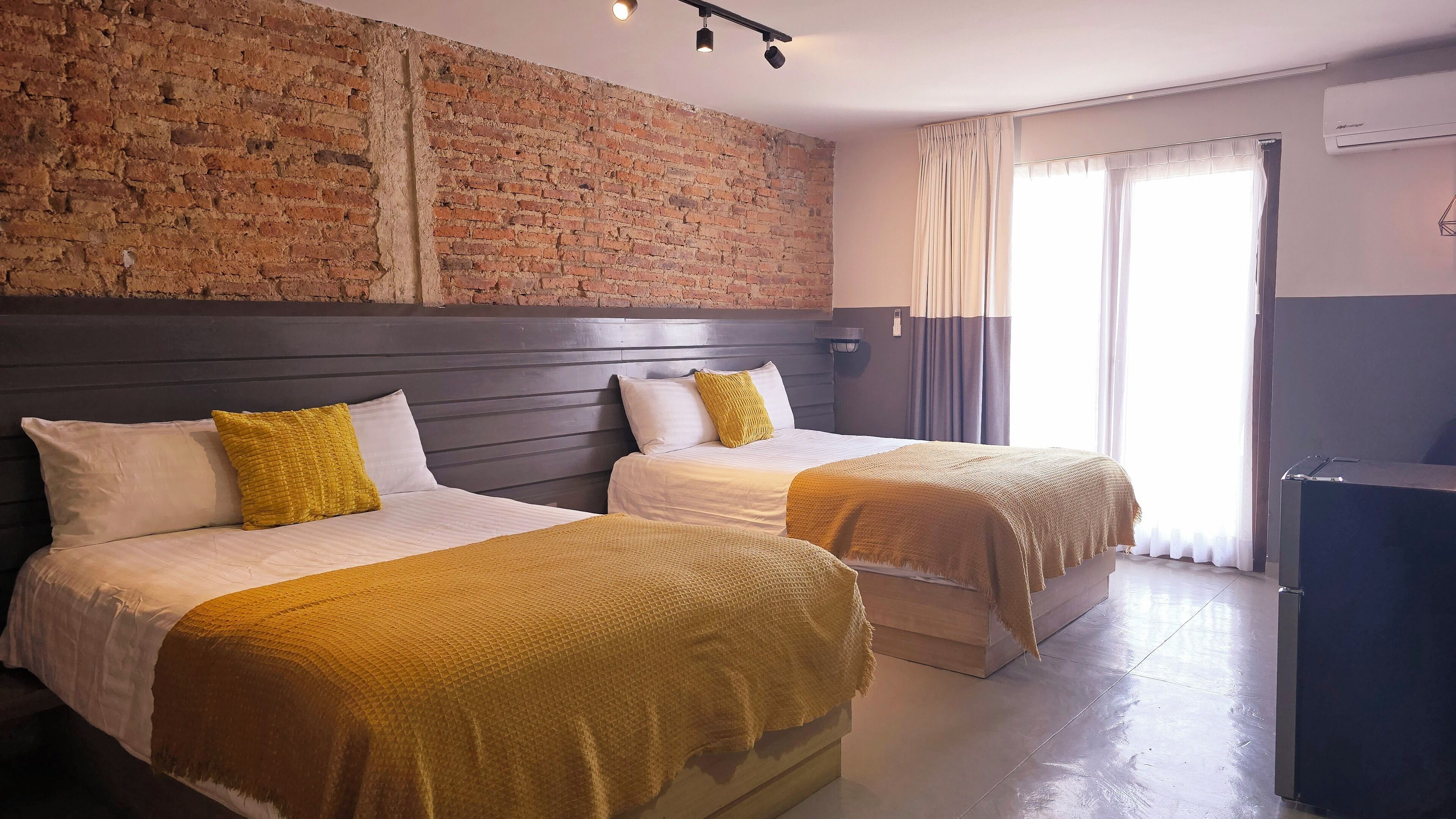 Een hotelkamer met twee bedden, een bakstenen muur, een groot raam met gordijnen en een airconditioningsunit.