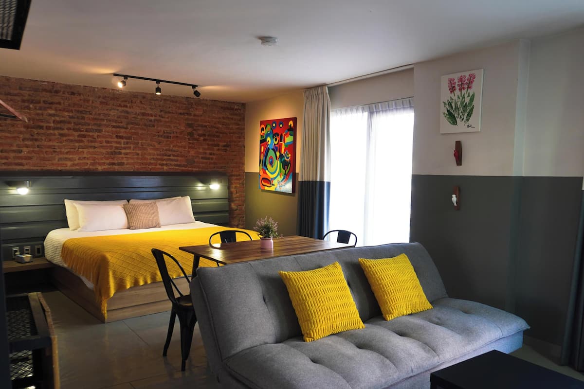 A modern hotel room with a bed, a sofa, a dining table, and a brick wall.