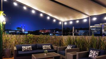 Un patio al aire libre con un sofá, sillas y una mesa, rodeado de plantas de bambú y vistas de la ciudad iluminada de noche.
