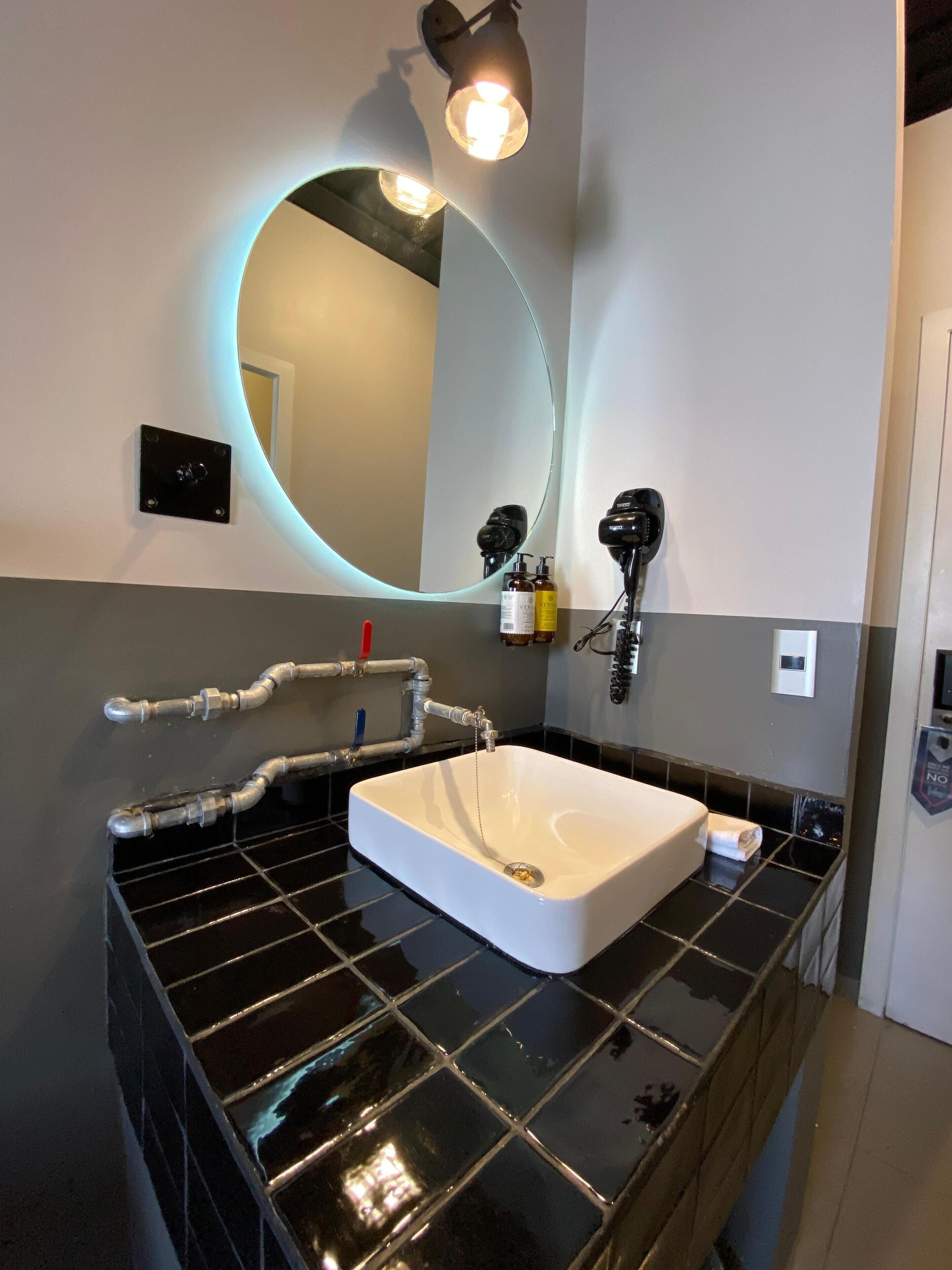 Un baño moderno con un lavamanos blanco, encimera con azulejos negros y un espejo grande con luz circular.