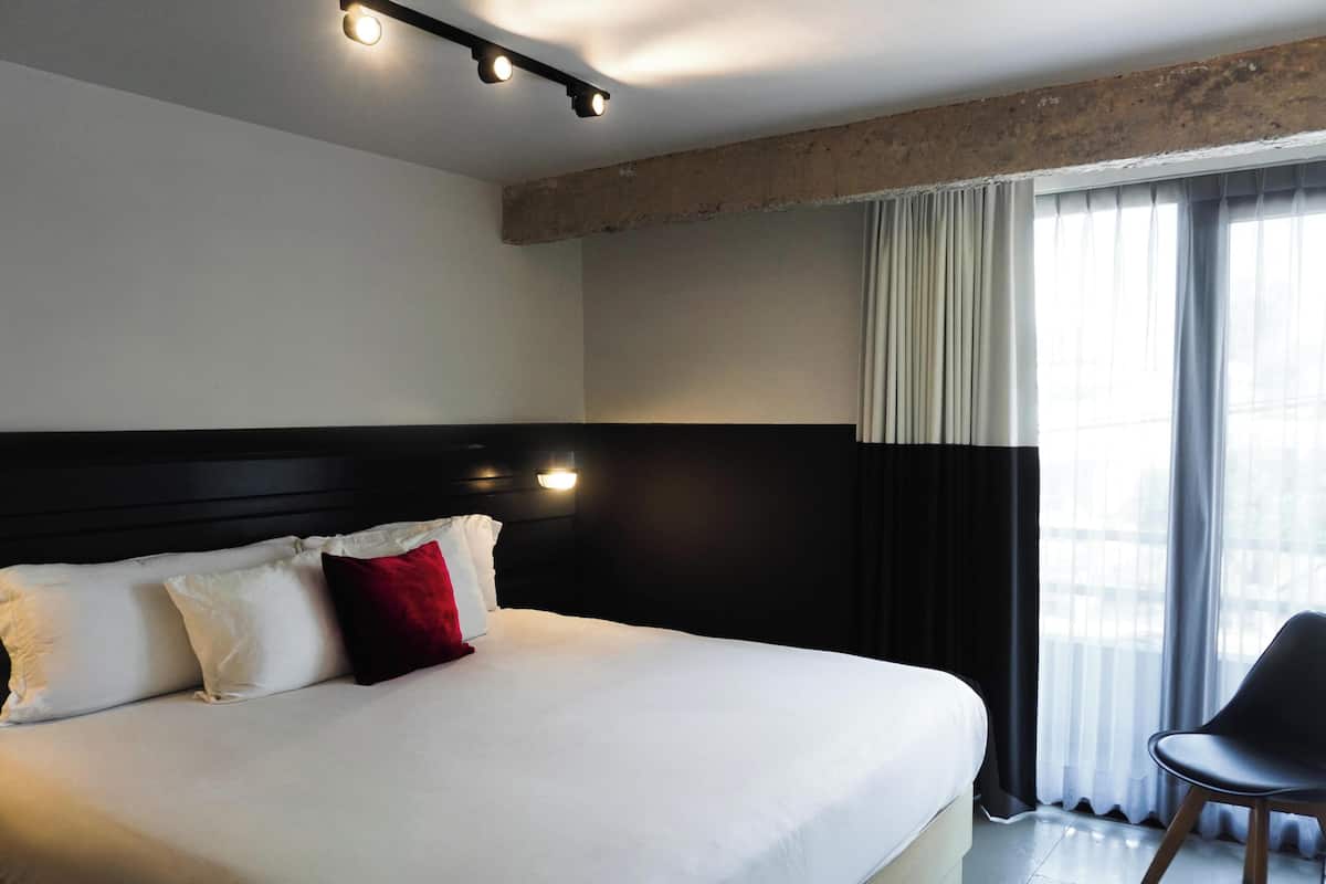 A hotel room with a large bed, a black headboard, a window with curtains, and a chair.