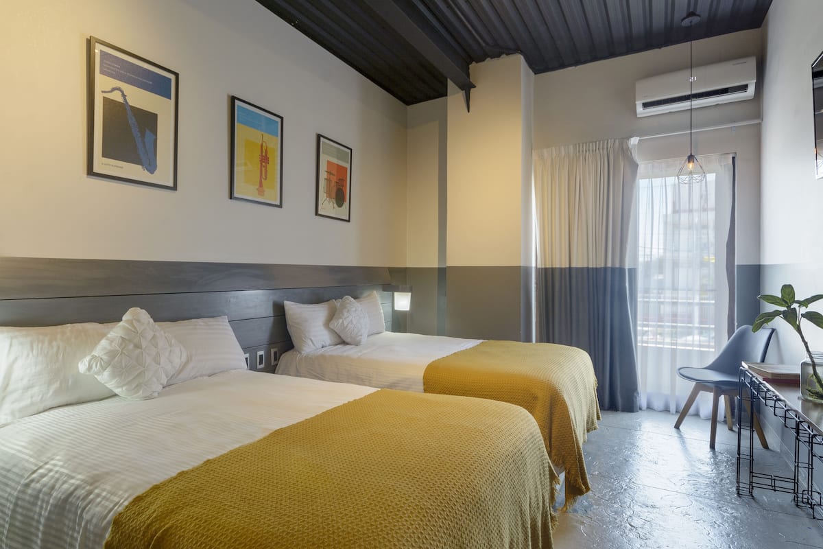 A hotel room with two beds, a desk with a chair, a potted plant, and artwork on the wall.