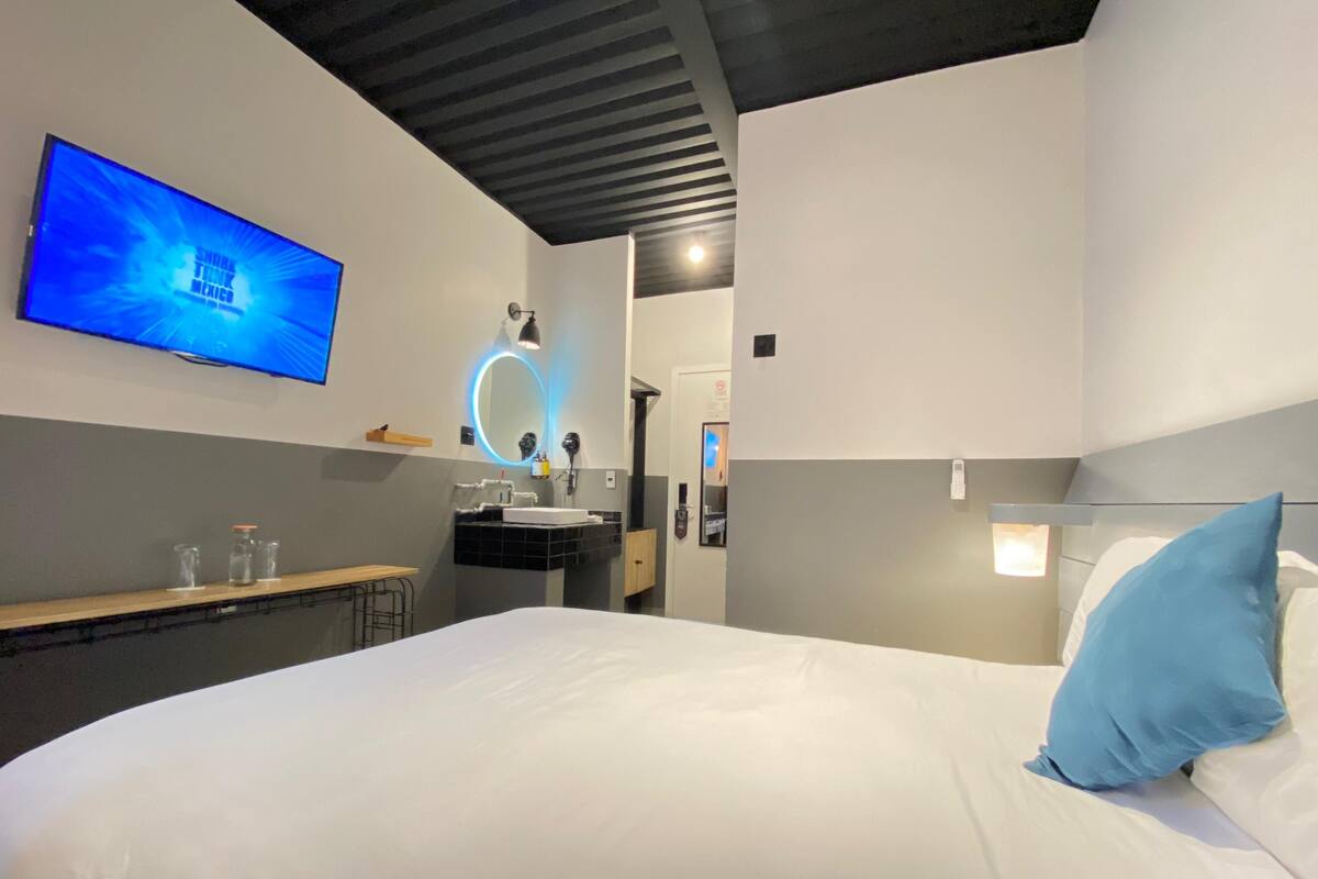 A modern hotel room with a large bed, a TV mounted on the wall, a bathroom with a mirror and sink, and a small table with glasses.