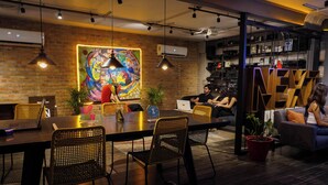 A modern meeting room with a brick wall, a colorful mural, and a mix of wicker chairs and sofas. - Hotel NEXT Guadalajara (Zapopan)
