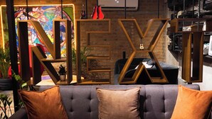 A modern interior with a large 'NEX' sign, a grey sofa with brown cushions, and a brick wall. - Hotel NEXT Guadalajara (Zapopan)