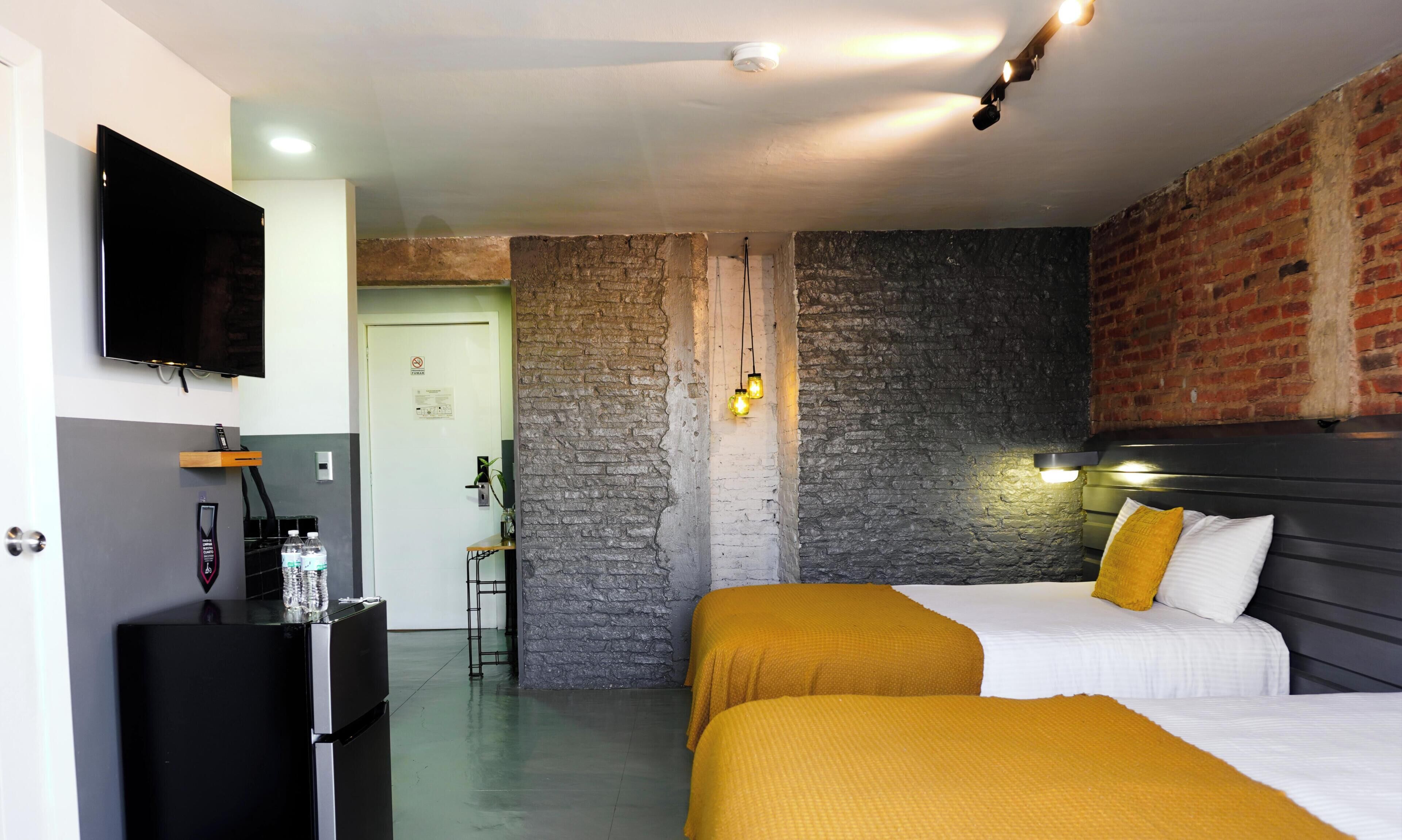 A hotel room with a bed, a desk with a refrigerator, a television, and a brick wall.