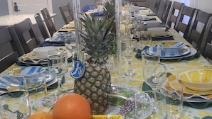 Une table à manger dressée pour un repas, avec un ananas en guise de centre de table, un panier d’oranges et un arrangement floral placé au-dessus.