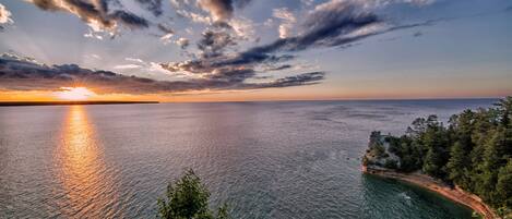 Un paisaje costero con atardecer sobre el agua, una costa rocosa y una zona boscosa.