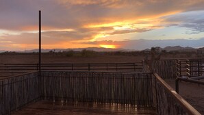 Eine Holzterrasse mit Metallgeländer, die auf eine Wüstenlandschaft mit Bergen in der Ferne und einem Sonnenuntergang blickt.