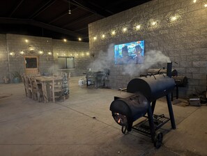 Eine Terrasse im Freien mit einer Steinwand, einem Holztisch und -stühlen, einem Grill und einem Fernseher, der ein Programm zeigt.