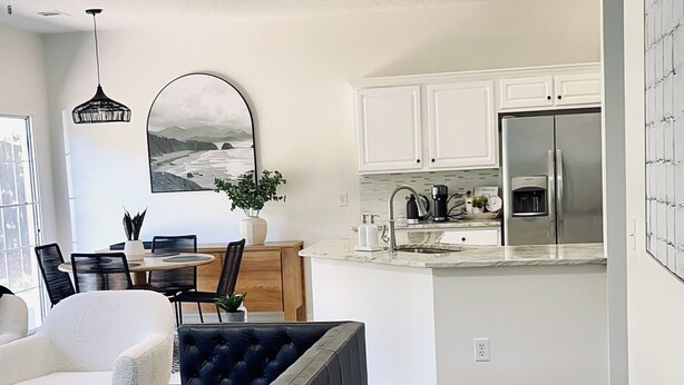 A modern living room with a black sofa, a dining area with a table and chairs, and a kitchen with white cabinets and stainless steel appliances.