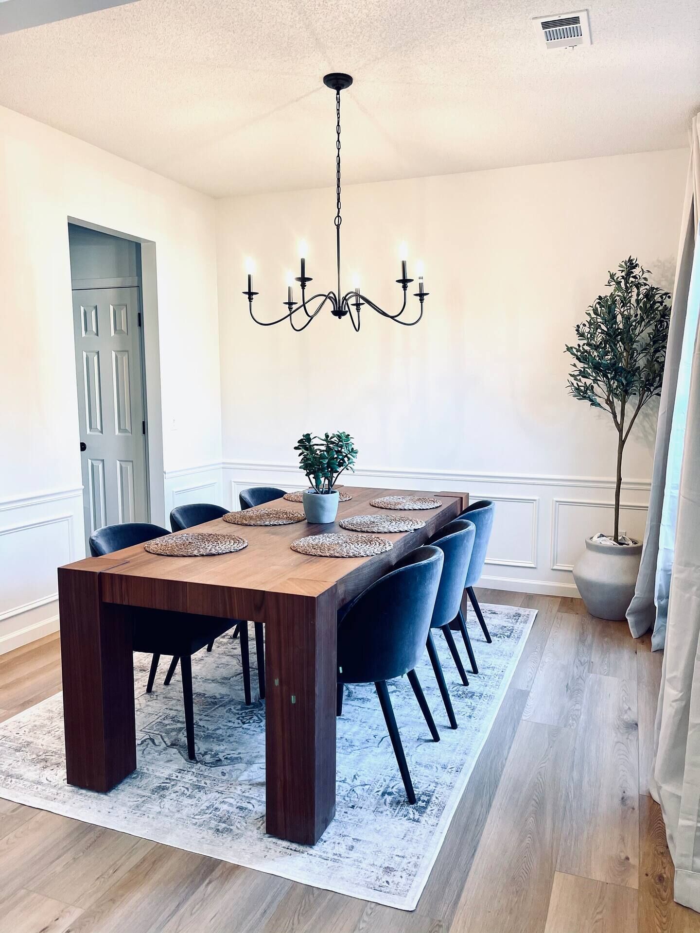 Une salle à manger avec une table en bois, six chaises, un lustre, une plante en pot et une porte.