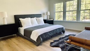 A modern bedroom with a large bed, a black headboard, two bedside lamps, a wooden floor, a fur rug, and a window with a view of trees.