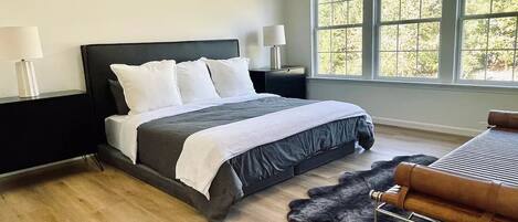 A modern bedroom with a large bed, a black headboard, two bedside lamps, a wooden floor, a fur rug, and a window with a view of trees.