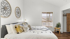 A bedroom with a large bed, two wall art pieces, a window with blinds, a small wooden stool, and a potted plant. - Updated condo with pools near golf courses (St. George)