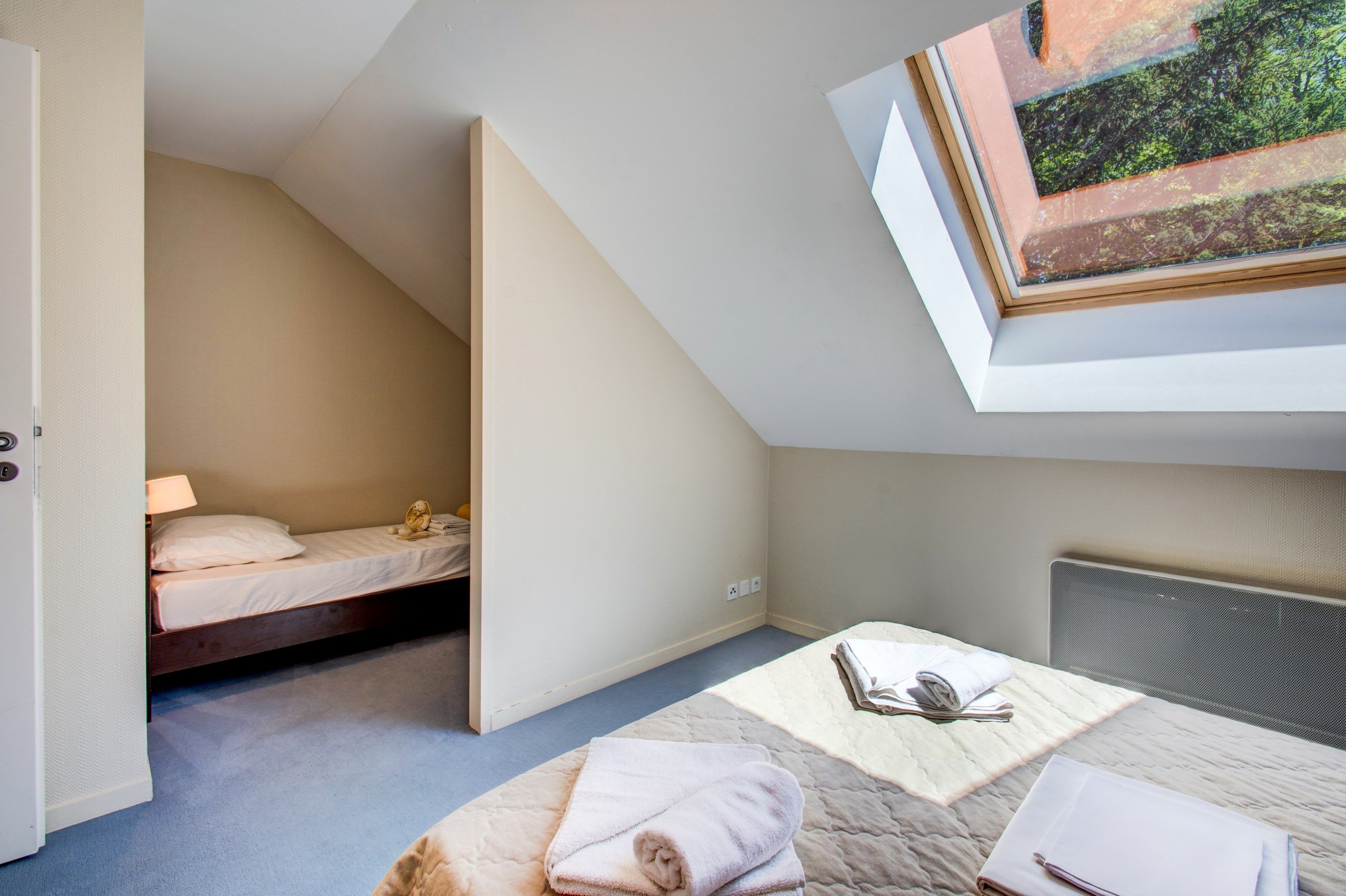 Une chambre avec une lucarne, un lit, une table de chevet avec une lampe, et une vue sur un jardin, visible à travers la lucarne.