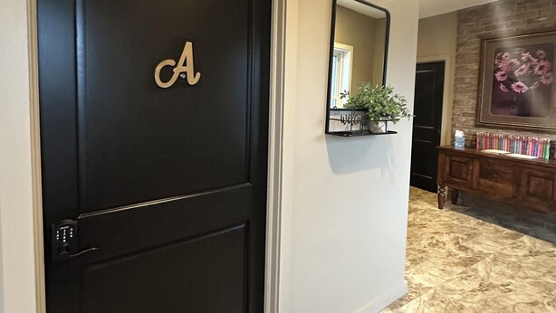 Una puerta negra con un gran monograma de la letra 'A', un felpudo, un espejo y un suelo de baldosas de piedra que conduce a una habitación con un banco de madera y un jarrón de flores.