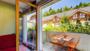 Un balcón con mesa y sillas, un sofá rojo, y vistas a edificios de madera y vegetación.