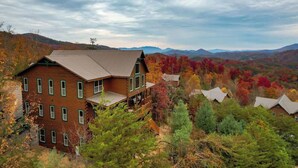 A large wooden building with multiple windows and a balcony, surrounded by autumn foliage and mountains in the background. - Luxury Cabin Awesome Mountain Views Sleeps 42 (Sevierville)