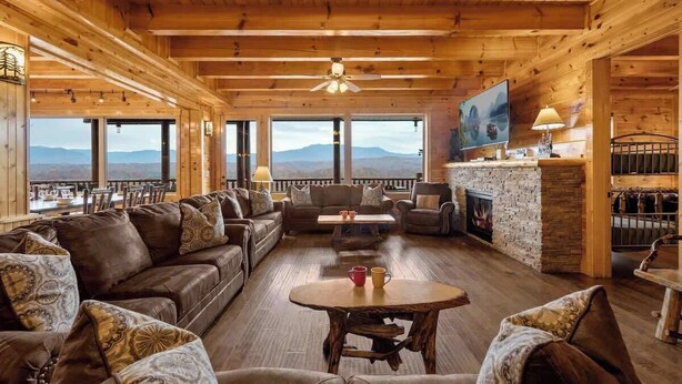 A wooden interior with a fireplace, seating area, and large windows offering mountain views. - Luxury Cabin Awesome Mountain Views Sleeps 42 (Sevierville)