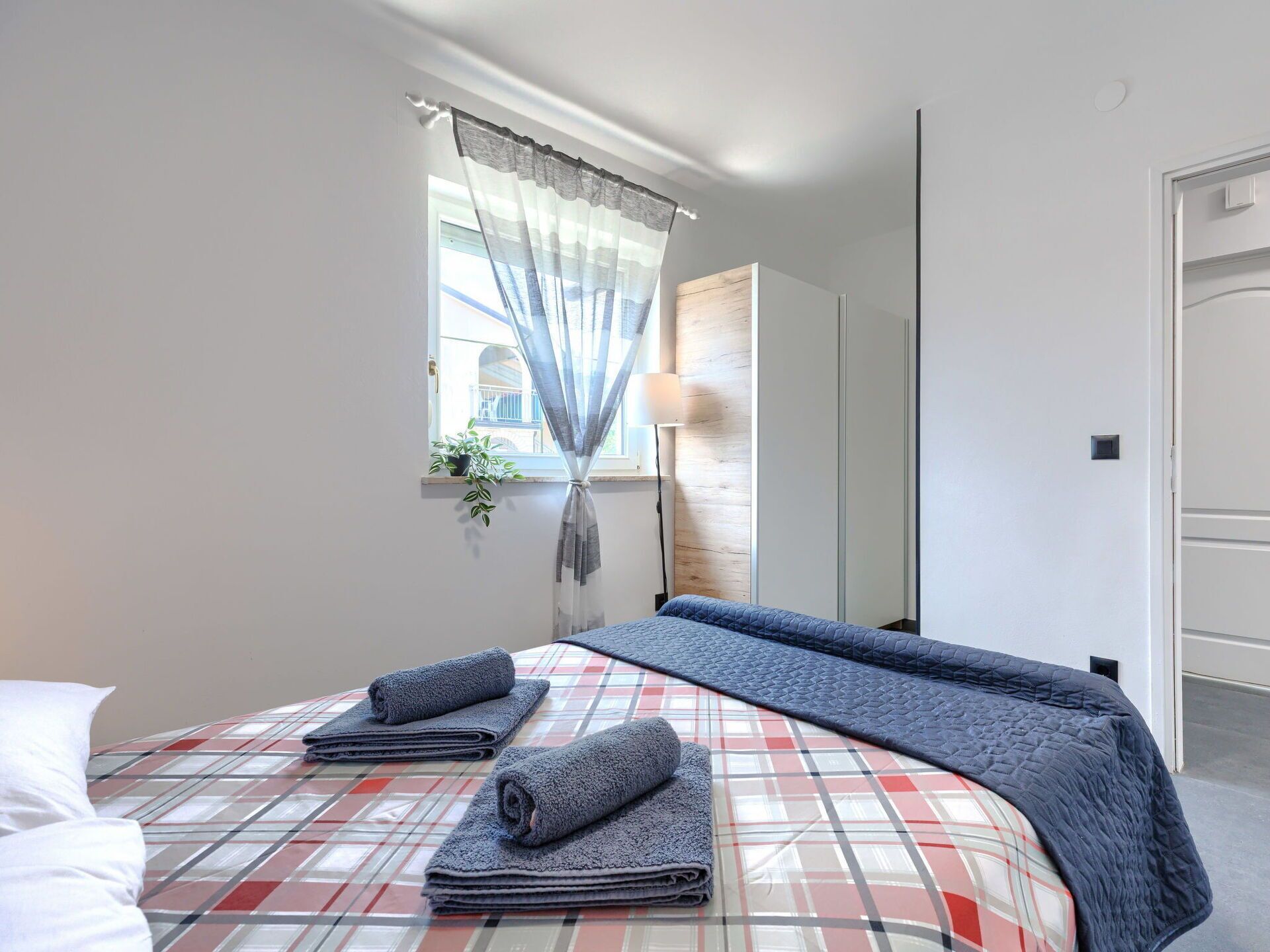 A bedroom with a bed, a window with sheer curtains, a bedside lamp, and a view of the outside.