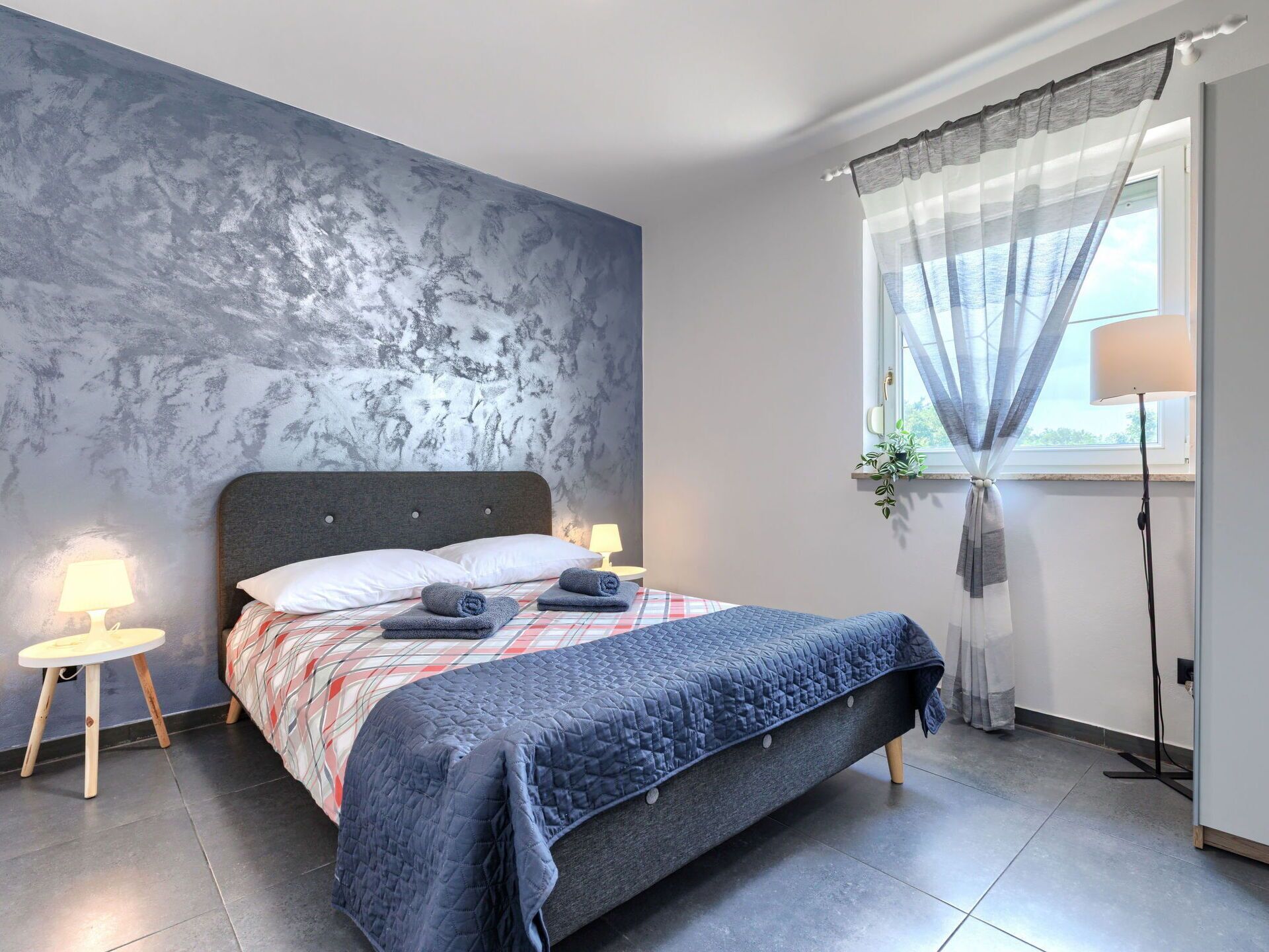 Quarto moderno com uma cama grande, abajures, uma janela com cortinas finas e uma parede texturizada.
